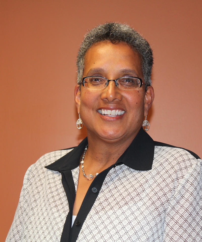 Charmaine is a Multiracial woman of Sri Lankan, Dutch, and Portuguese descent who believes most people see her as a Black woman. She has medium brown skin tone and her oval face has some freckles and darker age spots. Because of her high forehead the bridge of Charmaine's nose starts half way down her face. When not smiling, the edges of her mouth line up with the ends of her nostrils. Her teeth are tightly packed, so when she smiles her upper teeth form a connected line and her lower teeth are bunched up and a bit crooked. Charmaine's hair is mostly grey but has some black mixed in, and it forms tight curls about 2 inches around her head. She has brown eyes that appear behind her black rimmed glasses. 