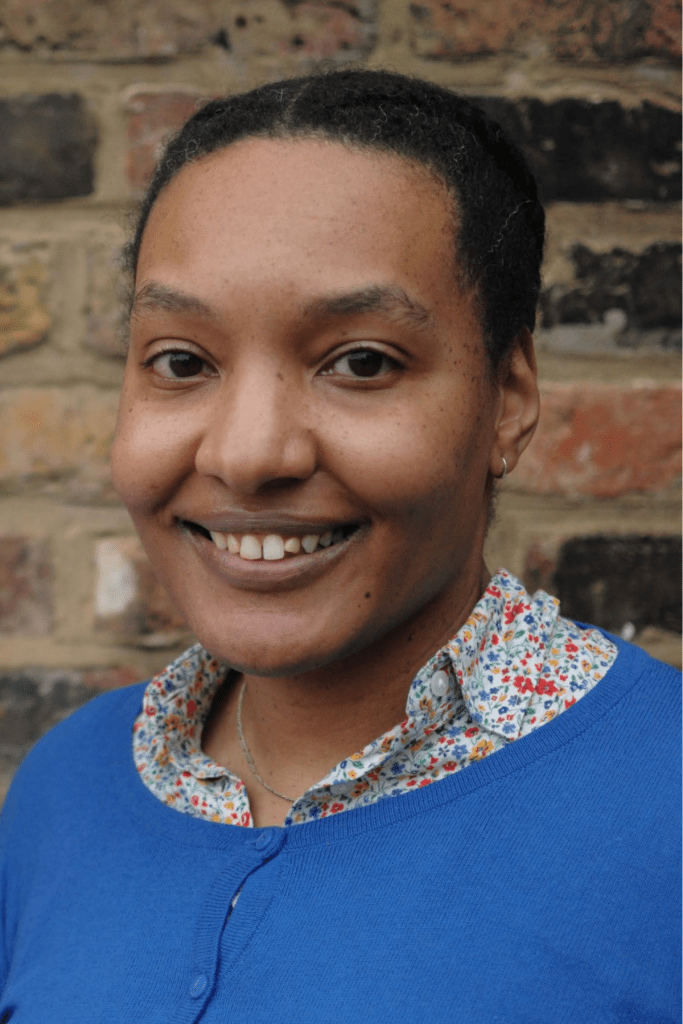 Chinelo L. Njaka is pictured outdoors from the waist up and standing in front of a sand-colored brick wall. Dr. Njaka wears a blue cardigan sweater over a white collared shirt with a ditsy floral print. She is smiling.