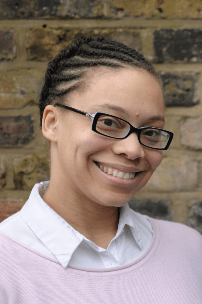 Jennifer Patrice Sims is pictured outdoors from the waist up and standing in front of a sand-colored brick wall. Dr. Sims wears a pink cardigan sweater over a white collared shirt and glasses. She is smiling.